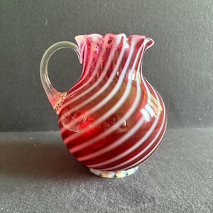 Puede incluir: Una pequeña jarra de cristal a rayas rojas y blancas con un asa transparente. La jarra tiene un borde con volantes y una base redonda. Las rayas son diagonales y espaciadas uniformemente. La jarra es probablemente vintage o antigua.