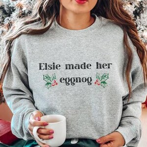 Puede incluir: Una sudadera gris con el texto "Elsie made her eggnog" y un pequeño diseño de acebo. La sudadera la lleva puesta una persona sentada en un sofá con un cojín de tiro a cuadros rojo y blanco.