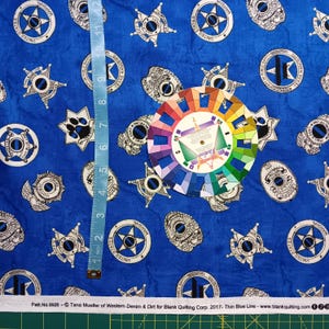 May include: Blue fabric featuring a repeating pattern of white and black law enforcement badges and paw prints. A light blue measuring tape shows 12 inches, and a circular color wheel is placed on the fabric. Text reads Patt.No.8928.