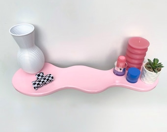 May include: A pink wavy shelf with a white vase, a black and white checkered hair clip, a pink candle, a small blue container, and a small white pot with a green succulent plant.