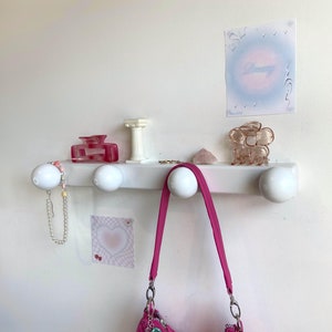 May include: White wall mounted shelf with four white ceramic hooks. A pink purse hangs from one hook. A pink hair clip, a pink and white beaded necklace, and a small white column are on the shelf. A pink heart sticker is on the wall below the shelf. A pink and white sticker with the word "Beauty" is on the wall above the shelf.