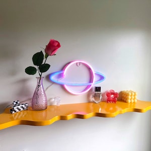 May include: A yellow, wavy shelf with a pink and blue neon Saturn, a pink rose in a clear glass vase, a small red flower, a clear glass bottle with a silver cap, and a small white object.