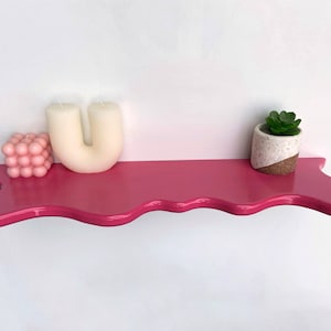 May include: A pink, wavy shelf with a white candle shaped like the letter U, a pink bubble-shaped object, and a small potted succulent plant.