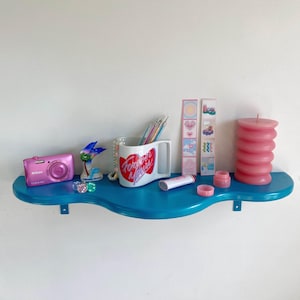May include: A blue shelf with a wavy edge, decorated with a pink camera, a white mug with the text "Forever Yours" in pink and red, a pink candle, and various other decorative items.