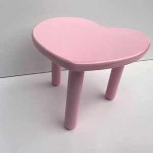Peut inclure: Un tabouret en forme de c&oelig;ur rose avec trois pieds. Le tabouret est en bois et a une finition lisse et brillante.