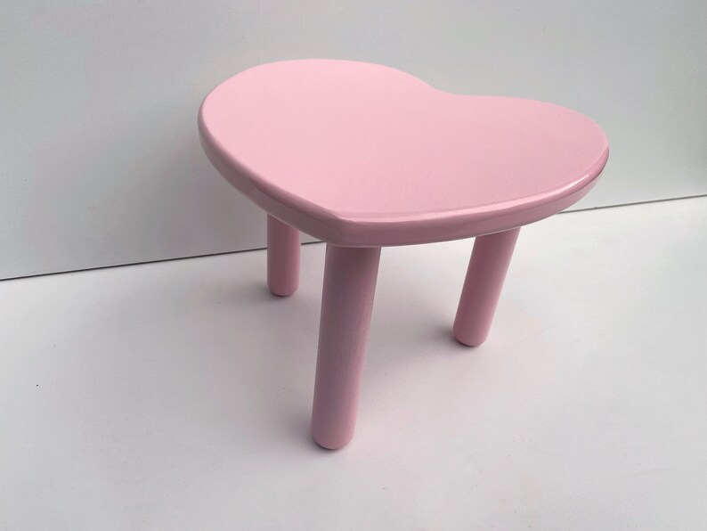 Peut inclure: Un tabouret en forme de c&oelig;ur rose avec trois pieds. Le tabouret est en bois et a une finition lisse et brillante.