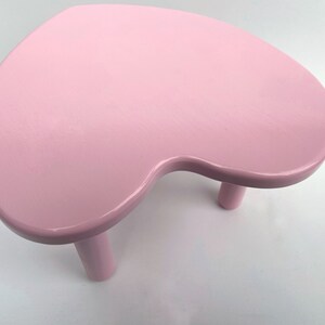 Peut inclure: Un tabouret en forme de c&oelig;ur rose avec trois pieds. Le tabouret est en bois et a une finition lisse et brillante.