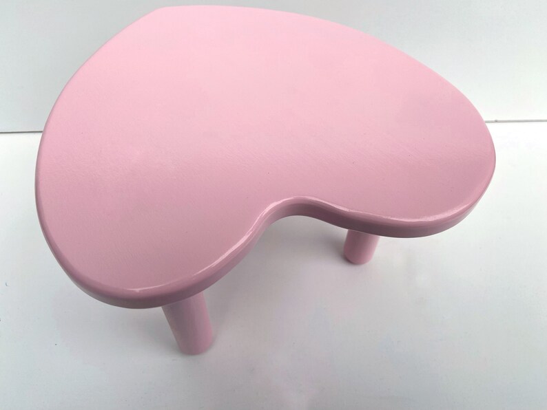 Peut inclure: Un tabouret en forme de c&oelig;ur rose avec trois pieds. Le tabouret est en bois et a une finition lisse et brillante.
