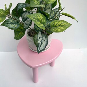 Peut inclure: Un support de plante en forme de c&oelig;ur rose avec trois pieds soutient un pot en c&eacute;ramique blanc avec une plante verte feuillue.