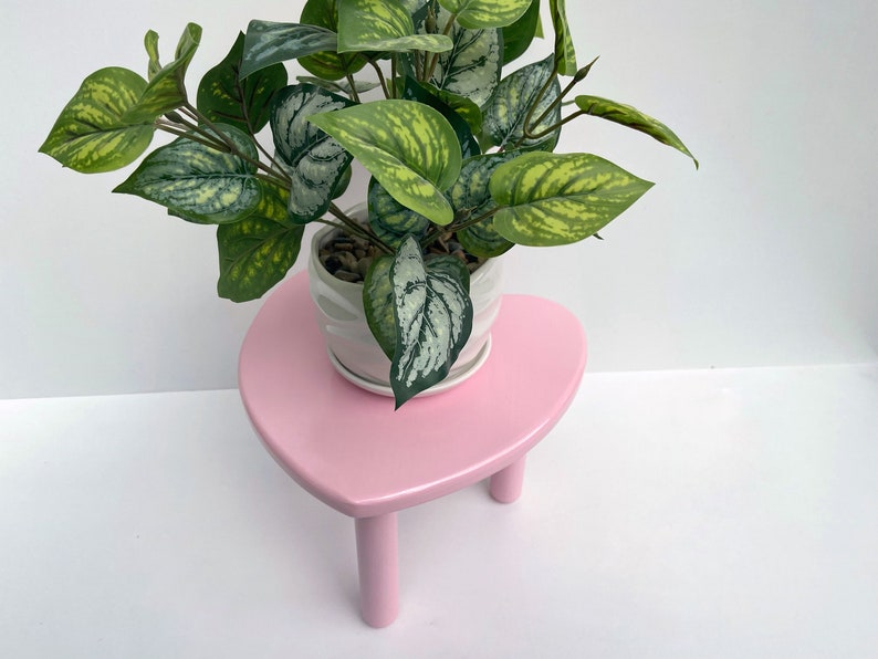 Peut inclure: Un support de plante en forme de c&oelig;ur rose avec trois pieds soutient un pot en c&eacute;ramique blanc avec une plante verte feuillue.