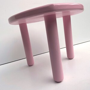 Peut inclure: Un tabouret en bois rose avec trois pieds. Le tabouret est peint d'une couleur rose clair et a une finition lisse et brillante.