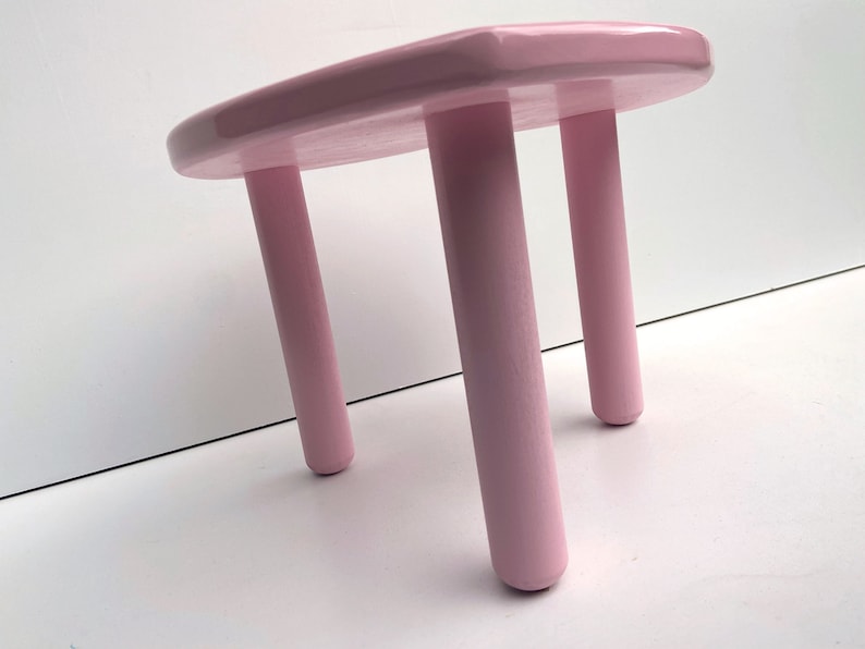 Peut inclure: Un tabouret en bois rose avec trois pieds. Le tabouret est peint d'une couleur rose clair et a une finition lisse et brillante.