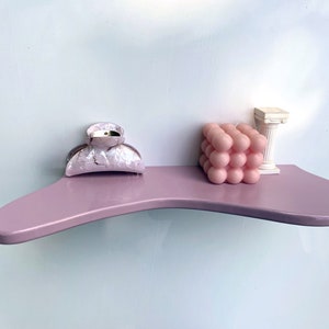 May include: A pink bubble candle, a white decorative column, and a pink and white hair clip are displayed on a light purple shelf.