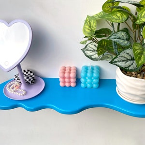 May include: A blue shelf with a wavy edge, featuring a heart-shaped mirror with a light, a pair of pink and blue bubble candles, and a small white planter with a green plant.