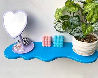 May include: A blue shelf with a wavy edge, featuring a heart-shaped mirror with a light, a pair of pink and blue bubble candles, and a small white planter with a green plant.