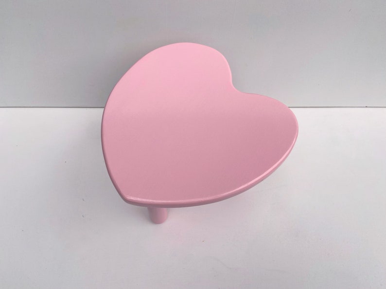 Peut inclure: Une table en forme de c&oelig;ur rose avec trois pieds. La table est en bois et a une finition lisse et brillante.