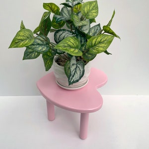 Peut inclure: Un support de plante en forme de c&oelig;ur rose avec trois pieds, soutenant un pot en c&eacute;ramique blanche avec une plante verte feuillue.