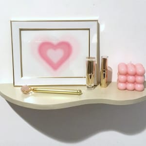 May include: A white shelf with a pink heart-shaped design in a white frame, a gold roller, two gold tubes, and a pink bubble candle.
