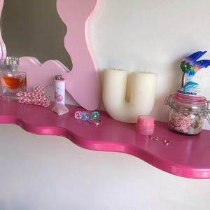 May include: A pink shelf with a wavy edge, decorated with a variety of items, including a bottle of perfume, a lighter, a candle shaped like the letter U, a small jar with a dolphin figurine on top, and a few rings.