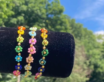 Puede incluir: Tres pulseras de cuentas coloridas con diseños de flores. Cada pulsera presenta un esquema de color diferente, incluyendo arcoíris, rosa y verde.