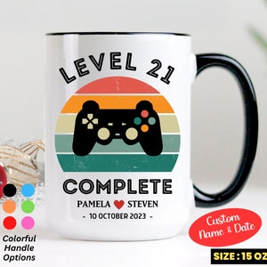 May include: White ceramic mug with a black handle. The mug features a retro-style video game controller graphic with the text "LEVEL 21 COMPLETE" and "PAMELA STEVEN - 10 OCTOBER 2023 -" printed on it. The mug is 15 ounces.