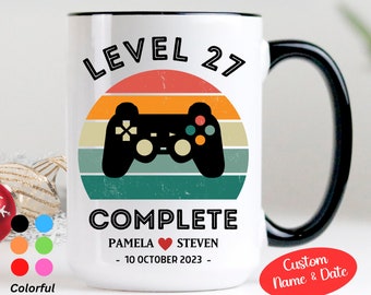 May include: White ceramic mug with a black handle. The mug has a retro-style graphic of a video game controller with the text "Level 27 Complete" and "Pamela ♥ Steven - 10 October 2023 -" printed on it. The mug is 15 ounces.