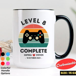 May include: White ceramic mug with a black handle. The mug features a retro-style video game controller graphic with a sunset background. The text "LEVEL 8 COMPLETE" is printed on the mug, along with the names "SOPHIA" and "STEVEN" and the date "10 OCTOBER 2023". The mug is 15 ounces.
