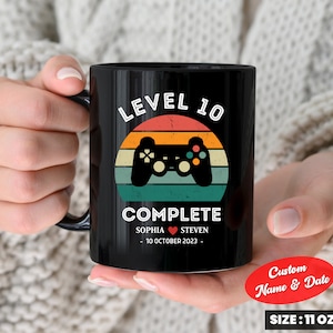 Pode incluir: Caneca de cerâmica preta com um design retro que apresenta um controlador de videogame e o texto "LEVEL 10 COMPLETE". A caneca também inclui os nomes "SOPHIA" e "STEVEN" e a data "10 DE OUTUBRO DE 2023". A caneca tem uma capacidade de 310 ml.