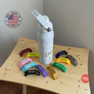 Puede incluir: Una botella de agua aislada blanca con un asa negra y una etiqueta que dice "Stanley Iceflow Bottle". La botella está rodeada de tapones de botella personalizados de colores con nombres como "Sophia", "Olivia", "Zania", "Noah", "Amelia", "Liam", "Elijah", "Emma", "Charlotte" y "James".