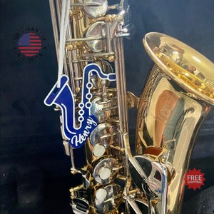 May include: A gold-colored saxophone with silver keys and a blue and white saxophone-shaped ornament that says "Henry". The ornament is hanging in front of the saxophone. The image also includes a "100% Made in America" sticker.