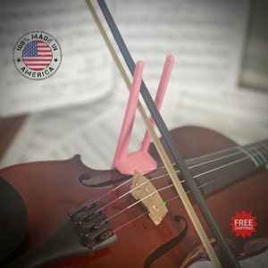 Pode incluir: Um violino castanho com um suporte rosa segurando dois arcos. Os arcos são pretos e castanhos. A imagem inclui o texto "100% MADE IN AMERICA" e uma pequena bandeira americana.