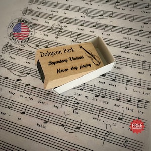 May include: A wooden stamp with the text "Dohyeon Park Legendary Violinist Never stop playing" and a drawing of a violin. The stamp is in a white box and is sitting on a sheet of music.