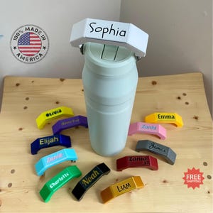 May include: An assortment of colourful personalised bottle stoppers with names such as Sophia, Elijah, Zania, Charlotte, Noah, Liam, Olivia, Amelia, Emma, and James. The stoppers are made of plastic and have a handle that can be used to open bottles.