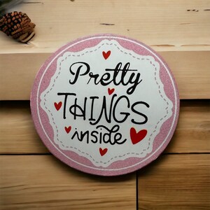 May include: Pink and white round sign with a pink and white patterned border. The sign says "Pretty Things Inside" in black lettering with red hearts.