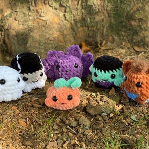 May include: A group of seven crocheted Halloween-themed plush toys. The toys are a ghost, a vampire, a bat, a Frankenstein, a werewolf, and two pumpkins. The toys are all sitting on a bed of brown leaves.