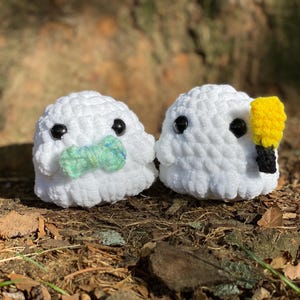 May include: Two white crocheted ghost plushies with black eyes. One ghost has a green and blue bow tie, and the other has a yellow beak.
