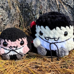 May include: Two handmade crochet vampire plushies. One is pink and black, the other is white and black. Both have large black eyes, fangs, and are sitting on a bed of dry grass.