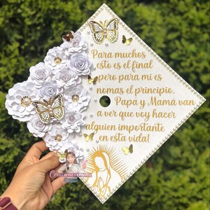 May include: A decorated graduation cap in white, adorned with white paper flowers, gold butterflies, and Spanish text. The cap features a pearl trim and a gold illustration of the Virgin Mary. The text says, "For many this is the end, but for me it's just the beginning."