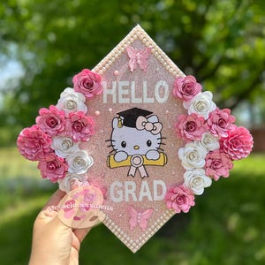 Peut inclure: Un bonnet de graduation rose décoré de fleurs en papier blanches et roses, une bannière "Hello Grad", un personnage Hello Kitty tenant un diplôme et des accents de perles.
