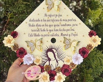 Puede incluir: Un birrete de graduación decorado con brillantina dorada, flores de papel en tonos burdeos, blanco y crema, y mariposas doradas. El birrete presenta una silueta de la Virgen María y el texto "Mi esfuerzo fue dedicado a los sacrificios de mis padres. Hubo dias en los que quise rendirme, pero recordé todo lo que han hecho por mi. Este titulo es para ustedes, Mamá y Papá."