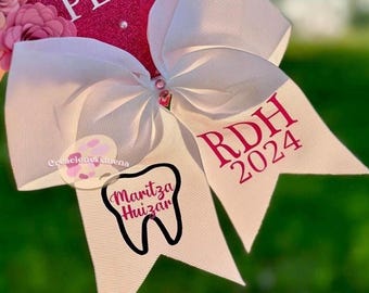 May include: White bow with pink text that says "RDH 2024" on one side and a pink tooth with the text "Maritza Huizar" on the other side.