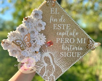 Puede incluir: Un gorro de graduación de brillantina dorada decorado con flores de papel blanco, mariposas y perlas. El gorro tiene una silueta blanca de la Virgen María y el texto "EL fin de ESTE capitulo PERO MI historia SIGUE".