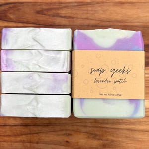May include: Four rectangular bars of soap with a marbled white and purple finish. One bar is wrapped in a tan paper band featuring the text soap geeks lavender patch and Net Wt. 6.5oz (184g) with a bubble pattern printed on the paper.