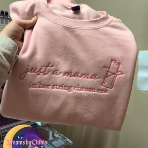 May include: A light pink sweatshirt with the embroidered text "just a mama in her string cheese era" in a matching pink thread. The design includes a string cheese graphic. The sweatshirt is held up by a hand.