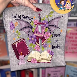 May include: A gray t-shirt with a fantasy-themed design. The design includes a sword, wings, books, flowers, and the text "lost in fantasy found in books." A butterfly and a key are also featured. The shirt is held up by a person.