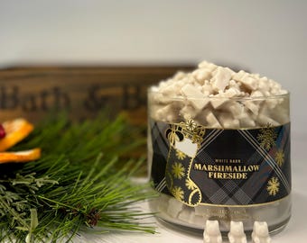 May include: A clear glass jar filled with small, off-white bear-shaped wax melts. The label features a black and blue plaid pattern, a gold stocking illustration, and the text White Barn Marshmallow Fireside. Three bear-shaped wax melts sit in front.