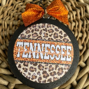 May include: A round, black, wooden ornament with a leopard print design and the word "Tennessee" in white letters. The ornament has an orange bow attached to it.