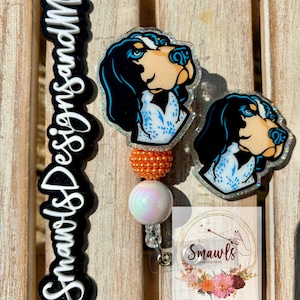 Może przedstawiać: Brelok do identyfikatora i pasująca odznaka z kreskówkowym psem w kolorach czarnym, niebieskim i brązowym. Brelok do identyfikatora ma pomarańczowe koraliki i perłową białą kulkę. Obraz zawiera również logo z tekstem "Smawls Designs and More".