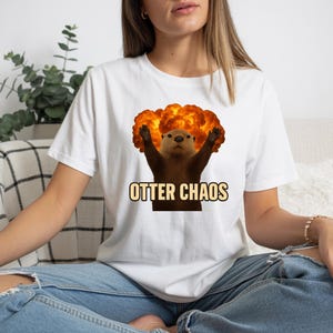 May include: White t-shirt featuring a cartoon otter with its arms raised in front of an orange explosion. The text "OTTER CHAOS" is printed below the otter in a yellow font. The shirt is worn by a person.