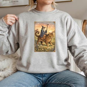 May include: A light gray sweatshirt featuring a whimsical illustration of a wizard riding a large tabby cat. The wizard wears a blue robe and hat, holding a staff. The cat walks through a field of flowers.